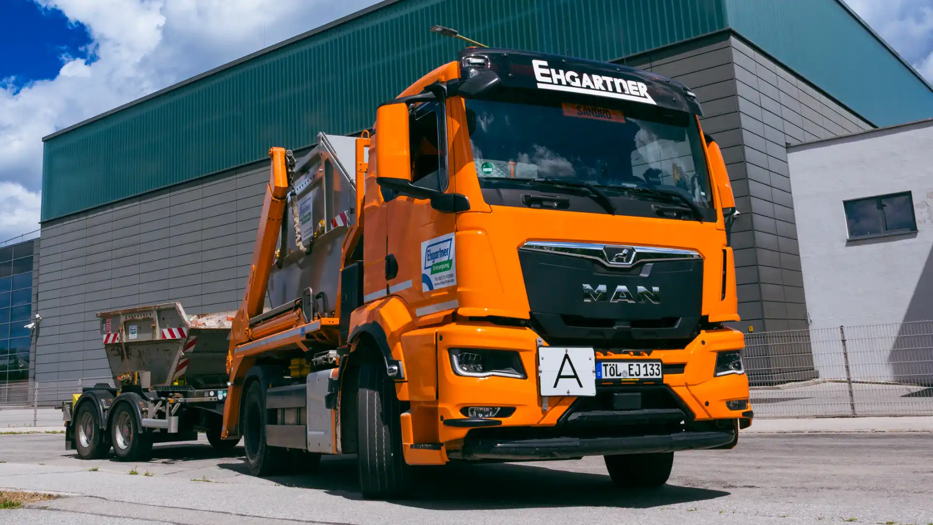 Ehgartner LKW mit Absetzcontainer und Anhänger auf einem Betriebshof vor einer Industriehalle.
